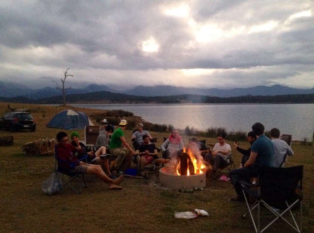 Campers sitting in folding chairs around a campfire by a lake as the sun sets behind the hills