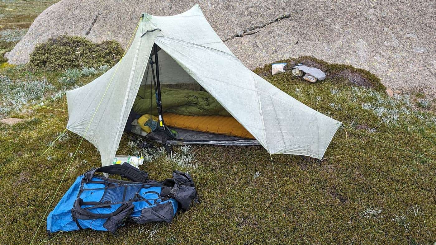 A lightweight white tent pitched on a grassy rock shelf with a backpack and gear in front