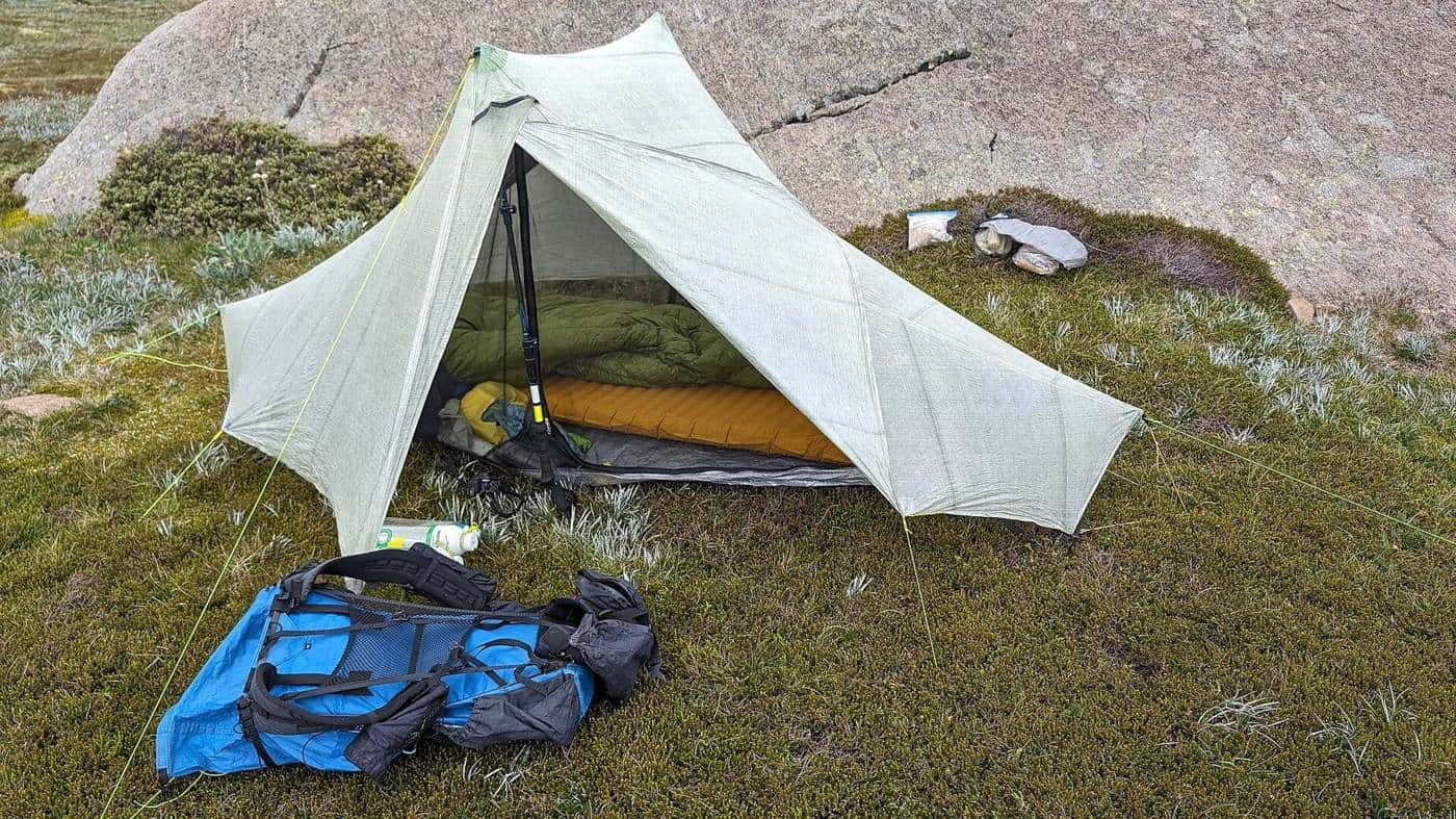 A lightweight white tent pitched on grass beside a granite slab, with a blue backpack and sleeping gear laid out in front